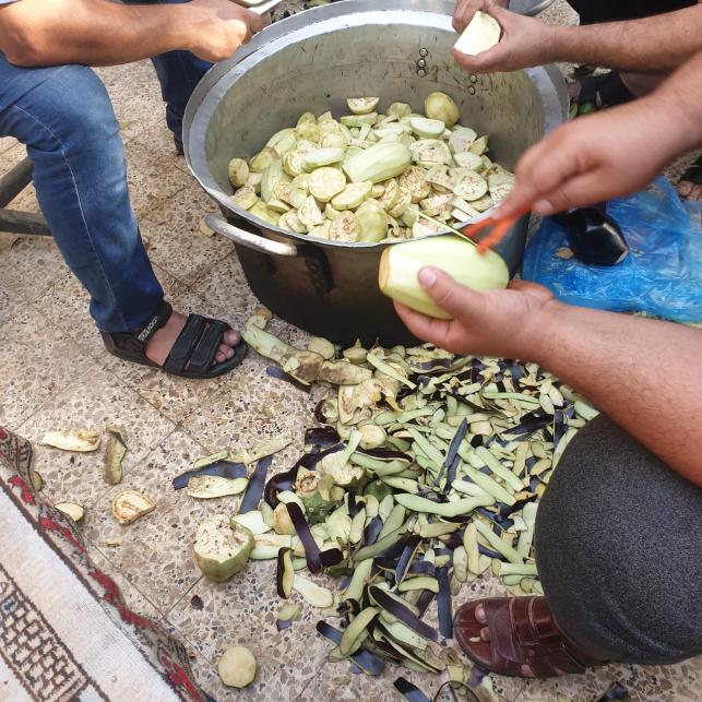 Men peeling egglant and putting pieces in large cooking pot.