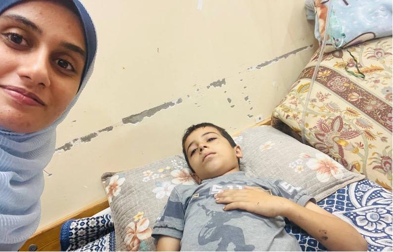 Boy lying in hospital bed with mother next to him, taking selfie.
