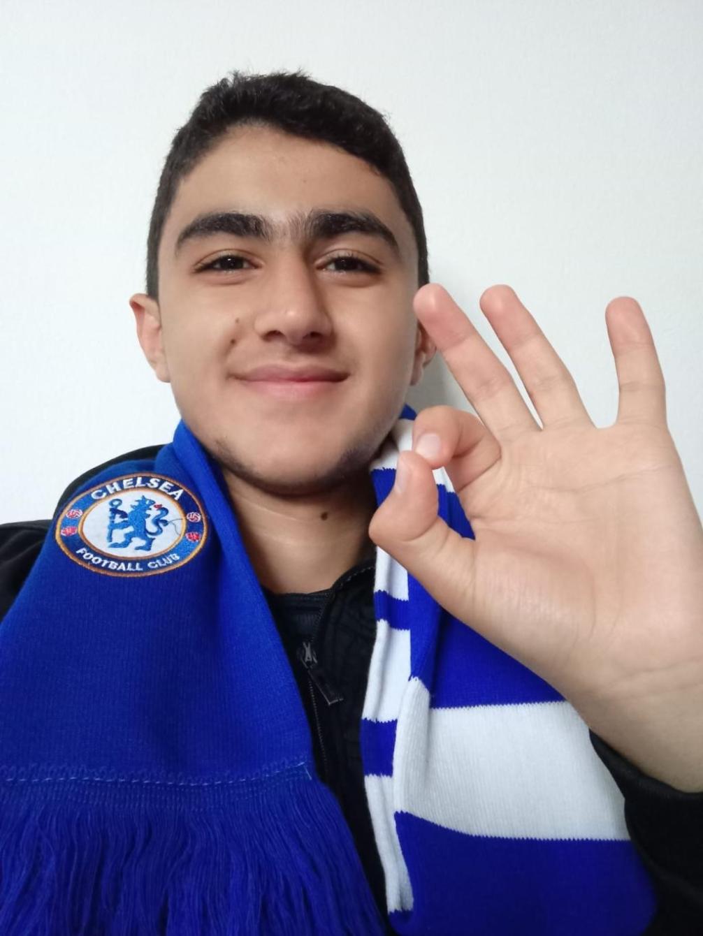 Abubaker Abed with Chelsea football club scarf, making okay sign.