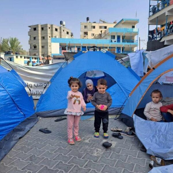 Two young children stand before a tent and their mother sits in the doorway of the tent which is in an encampment for displaced persons at a schoolyard in Gaza.