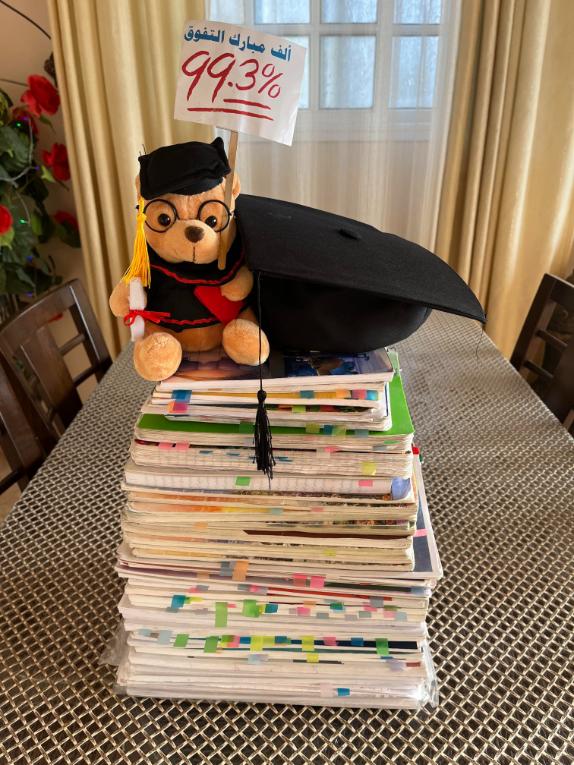 Teddy bear in graduation gown, holding flag that says "99.3%" and sitting on top of school notebooks.
