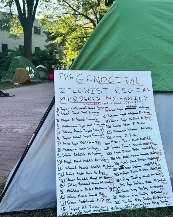 A large poster leaning against a tent. The poster text says: "The genocidal Zionist regime murdered my family" and it lists 19 people.
