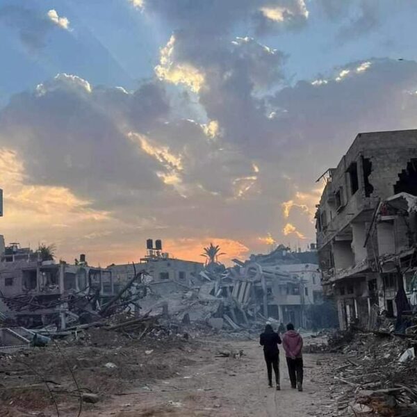 Two people in the distance walking on a street of destroyed buildings in Gaza. The sky is pink and blue.