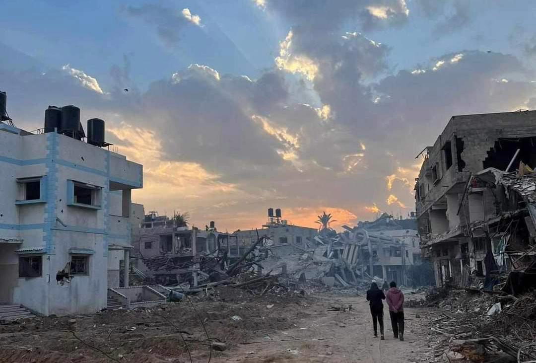Two people in the distance walking on a street of destroyed buildings in Gaza. The sky is pink and blue.