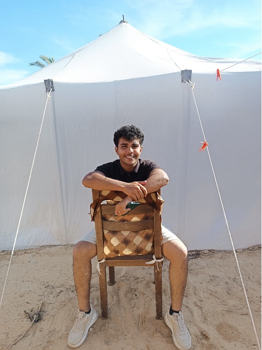 Young man sitting on chair, smiling, in front of a tent.