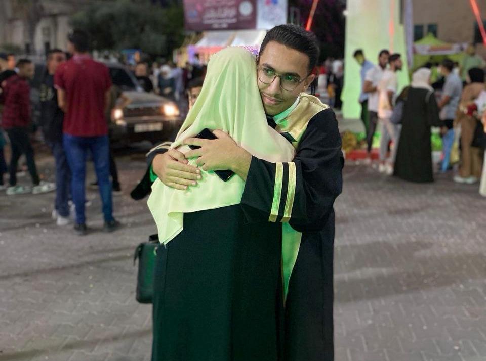 A young man in graduation gown embraced by his mother, outdoors around other people.